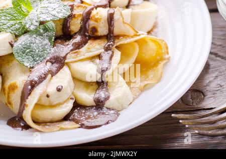 French crepes with chocolate sauce and banana in ceramic dish on wooden kitchen table Stock Photo