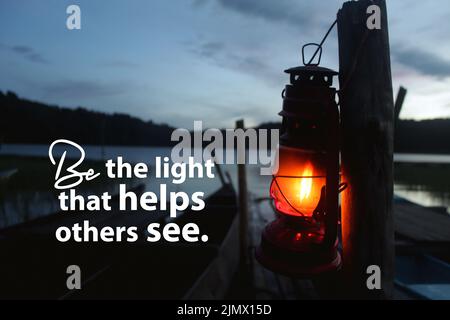 Life inspirational quote - Be the light that helps others see. With silhouette of a lantern hanging on wood in the lake. Stock Photo