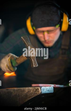 A man working blacksmith making a detail out of hot metal using a hammer Stock Photo