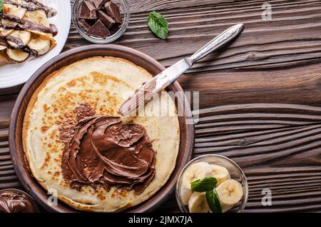 Flat lay French crepes with chocolate sauce and banana in clay bowl on wooden kitchen table Stock Photo