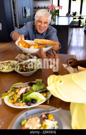 Smiling multiracial senior man giving breads to female friend while having lunch at dining table Stock Photo