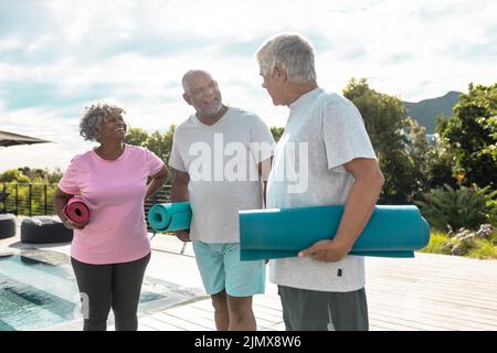 Happy multiracial senior friends with exercise mats talking while standing against sky in yard Stock Photo