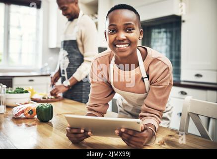 Looking for the perfect recipe. Cropped portrait of an attractive young woman using a tablet in the kitchen while her husband cooks in the background. Stock Photo