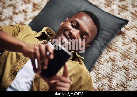 Somebody just got a text from their crush. High angle shot of a handsome young man smiling while using a smartphone while lying on the floor in his Stock Photo