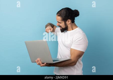 Portrait of aggressive man with beard wearing white T-shirt working on laptop computer, has mistake, wants to bang her fist on the screen. Indoor studio shot isolated on blue background. Stock Photo