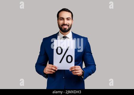 Positive smiling bearded man holding showing paper with percent sign inscription, looking at camera, wearing official style suit. Indoor studio shot isolated on gray background. Stock Photo