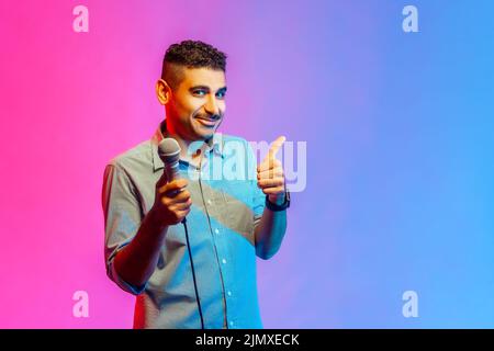 Portrait of attractive man in shirt posing with microphone in hands, offers mic, journalist asking questions, showing thumb up. Indoor studio shot isolated on colorful neon light background. Stock Photo