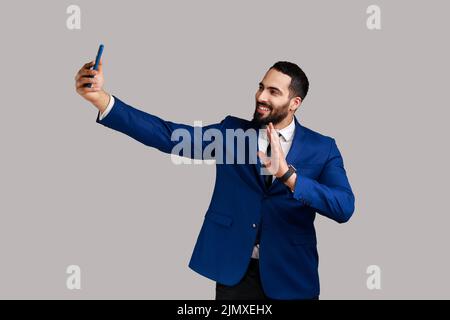 Man making selfie on smartphone camera, blogger communicating, recording video for followers, waving hand, saying hello, wearing official style suit. Indoor studio shot isolated on gray background. Stock Photo