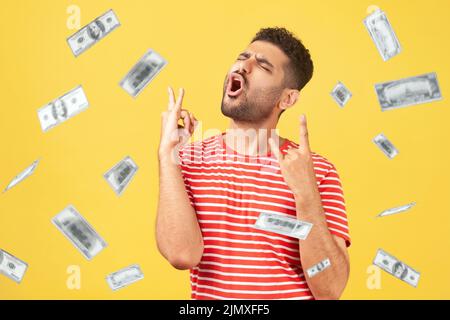 Money rain, winner and rich. Ecstatic happy celebrating motivated man standing with rock sign and shouting for joy, winner excited for success. dollars falling. indoor isolated on yellow background Stock Photo