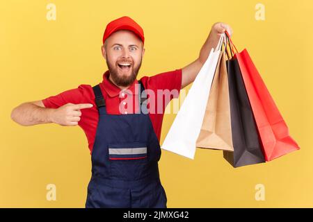 Portrait of cheerful positive delivery man in uniform pointing finger at shopping bags in his hand, fast delivery from store, service industry. Indoor studio shot isolated on yellow background. Stock Photo