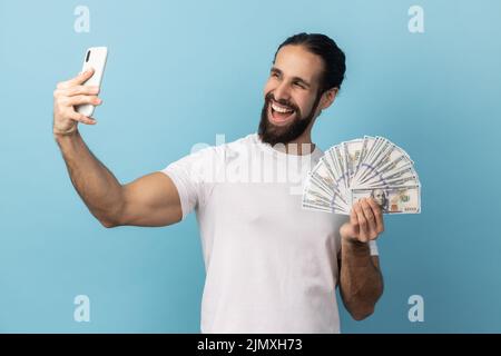 Portrait of happy smiling handsome man with beard wearing white T-shirt taking selfie on cellphone with fan of dollar banknotes in hands. Indoor studio shot isolated on blue background. Stock Photo