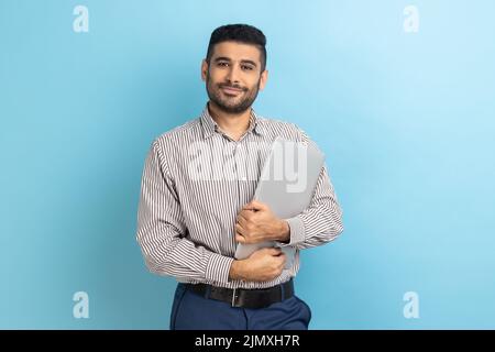 Portrait of smiling businessman standing holding closed laptop or folder, looking at camera with positive expression, wearing striped shirt. Indoor studio shot isolated on blue background. Stock Photo