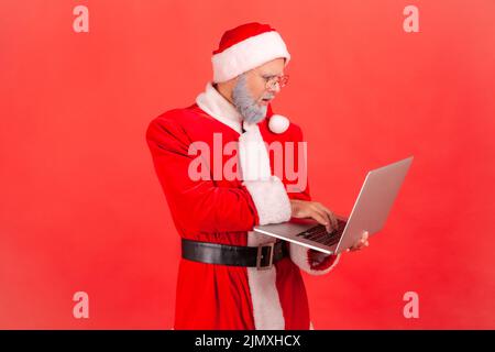 Side view of serious concentrated man elderly man with gray beard wearing santa claus costume working on laptop computer, thinking about new project. Indoor studio shot isolated on red background. Stock Photo