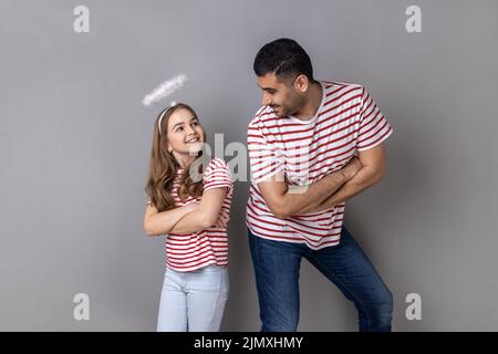 Portrait of smiling joyful father and daughter in striped T-shirts standing with folded hands and looking at each other, family expressing happiness. Indoor studio shot isolated on gray background. Stock Photo