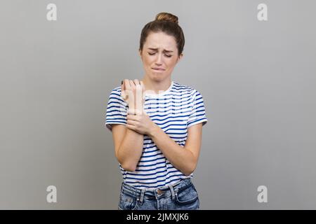 Portrait of unhappy sick woman wearing striped T-shirt touching painful hand, suffering trauma, sprain wrist, feeling ache of carpal tunnel syndrome. Indoor studio shot isolated on gray background. Stock Photo