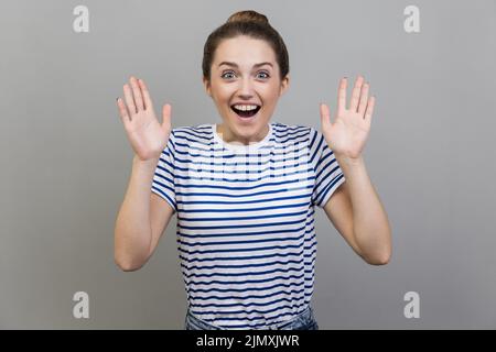 Portrait of extremely happy positive excited woman wearing striped T-shirt standing with raised arms, looking at camera, being pleasantly surprised. Indoor studio shot isolated on gray background. Stock Photo