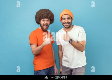 Well done, excellent job. Two young adult men showing thumbs up, gesturing like to camera and smiling broadly, expressing approval, positive feedback. Indoor studio shot isolated on blue background. Stock Photo