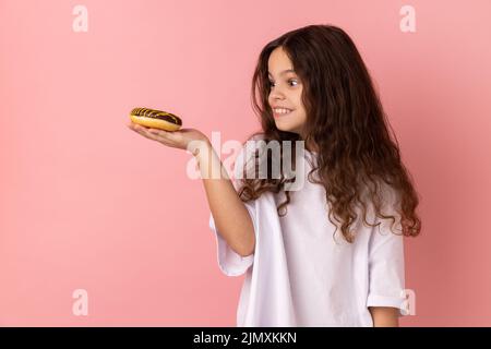 Portrait of hungry little girl wearing white T-shirt looking at donut with excited expression and want to eat confectionary, junk food. Indoor studio shot isolated on pink background. Stock Photo