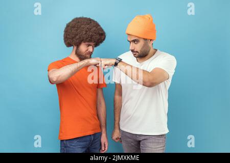 Portrait of two serious young adult hipster men standing and looking at each other with aggressive look and with bump gesture, fighting. Indoor studio shot isolated on blue background. Stock Photo