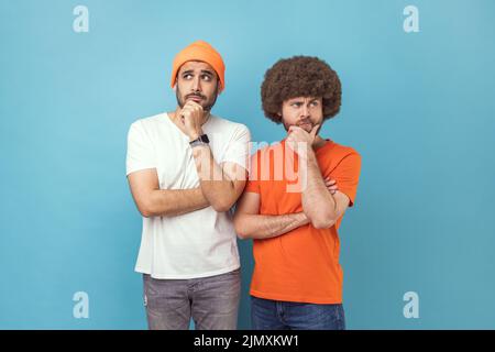 Portrait of two young adult hipster men holding chin with hands, need to think, planning strategy, thinking over ideas for startup. Indoor studio shot isolated on blue background. Stock Photo