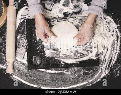 Two female hands knead the dough from white wheat flour on a black table, top view Stock Photo