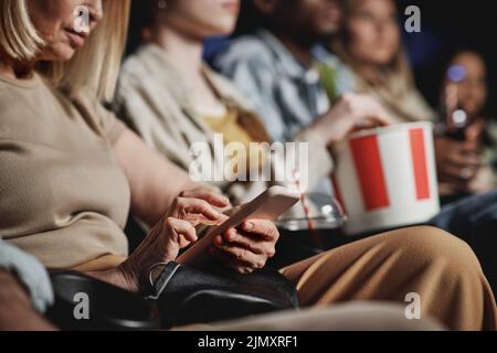 Selective focus of mature Caucasian woman surfing Internet on smartphone while watching movie at cinema Stock Photo
