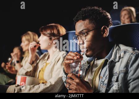 Selective focus shot of stylish young Black guy wearing eyeglasses drinking cola while watching movie at cinema Stock Photo