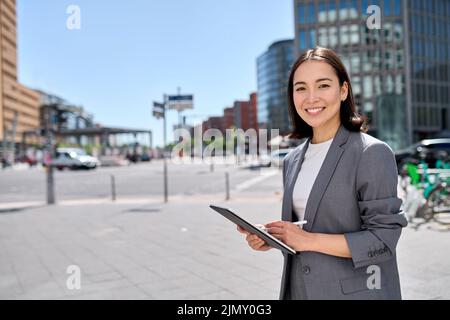 Smiling young Asian business woman standing on city street using digital tablet. Stock Photo