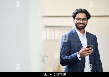 Smiling indian business man using phone standing in office, portrait. Stock Photo