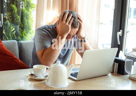 Upset business man sitting at table with laptop in cozy cafe and holding hands on head. Implementation of modern technologies into lifestyle. Remote Stock Photo