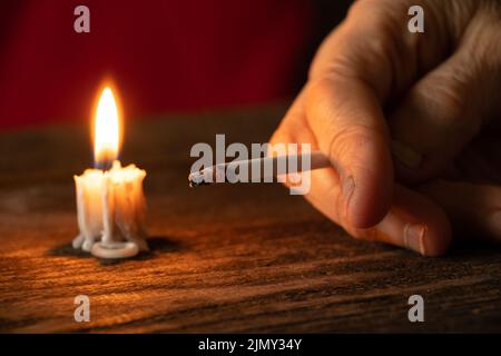 old woman holding a cigarette by candlelight in the dark at home at the table, smoking at home in the dark, nicotine and bad habit, cigarettes and can Stock Photo