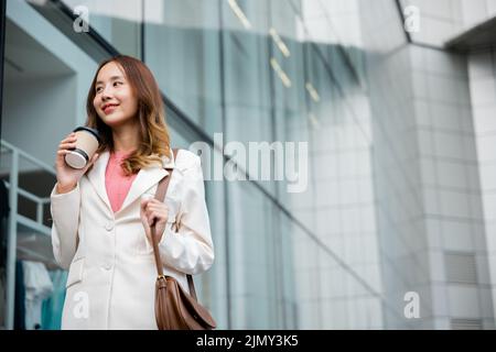 Asian businesswoman holding coffee cup takeaway going to work she walking near her office building, Portrait smiling business woman hold paper cup of Stock Photo