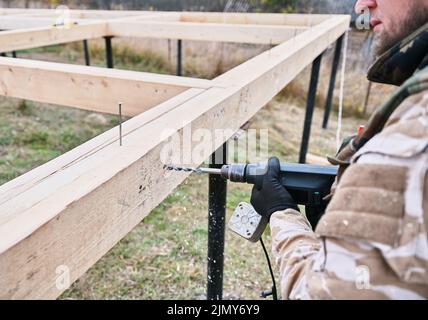 Man building wooden house on pile foundation. Close up of male worker drilling hole by electric drill in wooden frame of future house on construction site. Carpentry concept. Stock Photo