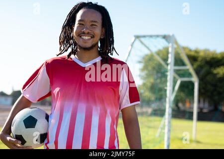 Smiling biracial male player with dreadlocks wearing red jersey with ball standing against clear sky Stock Photo