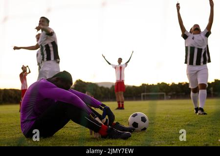 Multiracial sad goalkeeper sitting on grassy land and players with arms raised celebrating victory Stock Photo