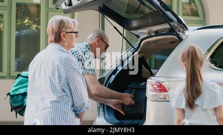 Positive family going on field trip while loading luggage inside car trunk. Grandparents, mother and daughter getting ready for summer citybreak while putting baggage and trolleys inside vehicle. Stock Photo