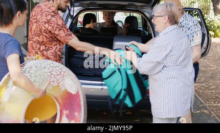 Happy people loading vehicle with luggage and trolleys while going on weekend citybreak together. Big family packing voyage luggage in car trunk while getting ready for summer field trip. Stock Photo