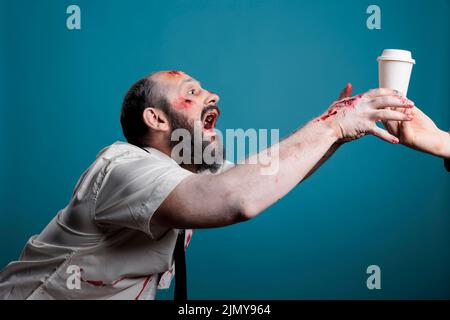 Aggressive zombie wishing to drink coffee cup in studio, going after beverage and wanting to eat carbon. Spooky undead scary devil chasing hand with drink, acting dangerous and creepy. Stock Photo