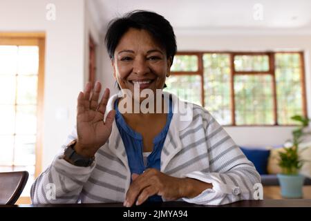 Portrait of smiling biracial mature woman with short hair waving hand while sitting at home Stock Photo