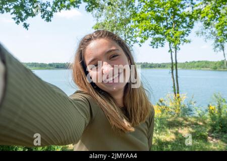 Beautiful cheerful young woman having a good time at the forest lakeside on a lovely day, taking a selfie, smiling Stock Photo