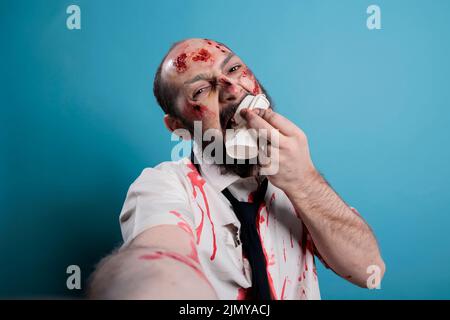 Halloween monster eating carton cup on camera, taking picture over blue background. Dangerous looking evil zombie biting on coffee cup to act frightening and scary, creepy deadly devil. Stock Photo