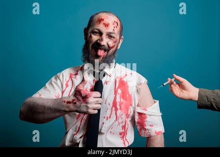 Scary zombie approving with covid 19 vaccine for protection, using syringe over blue background. Undead evil monster giving thumbs up like about needle to show approval and like, aggressive corpse. Stock Photo