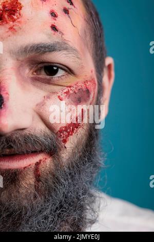 Halloween zombie having bloody wounds and scars on half of face, looking aggressive and evil in front of studio camera. Dangerous wounded brain eating monster acting scary. Close up. Stock Photo