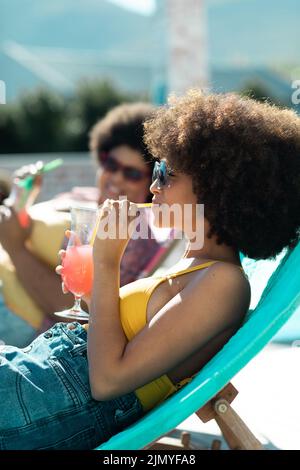 Biracial woman in sunglasses with afro hair drinking cocktail while enjoying pool party with friend Stock Photo