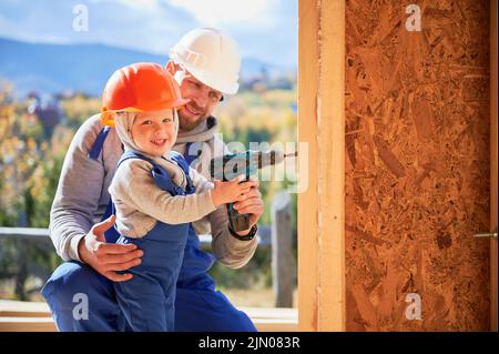 Father with toddler son building wooden frame house. Male builder and kid using screwdriver on construction site, wearing helmet and blue overalls on sunny day. Carpentry and family concept. Stock Photo
