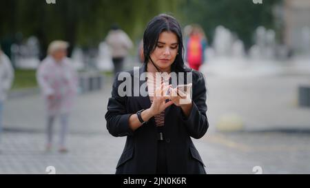Close-up businesswoman stands outdoors in city near roadway holding mobile phone looking around focused hispanic girl trying find building using Stock Photo