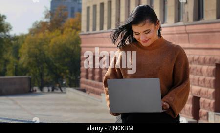 Young businesswoman freelancer student sitting outdoors reading email on laptop smiling laughing good news feels happy enjoys process surf web on Stock Photo
