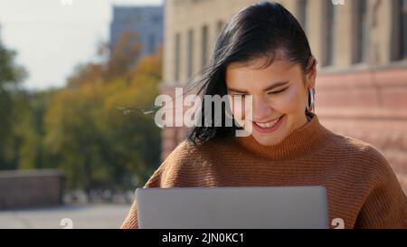 Young businesswoman freelancer student sitting outdoors reading email on laptop smiling laughing good news feels happy enjoys process surf web on Stock Photo
