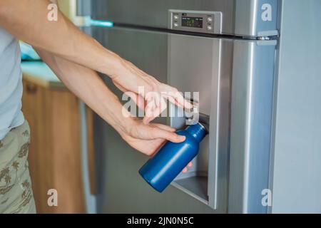 Male hand is pouring cold water and ice cubes in a metal bottle from dispenser of home fridge Stock Photo