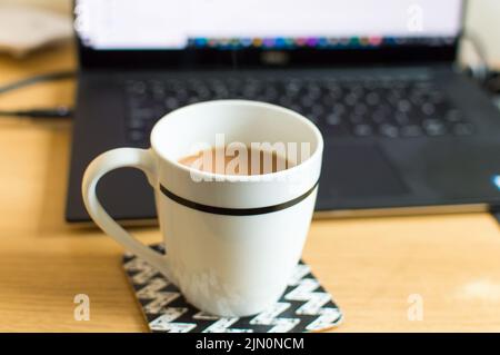 A cup of tea on a coaster next to a laptop workstation Stock Photo